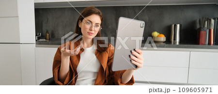 Portrait of young woman working from home, sitting in kitchen and talking with team, has online meeting, using digital tablet to join webinar, having conversation 109605971