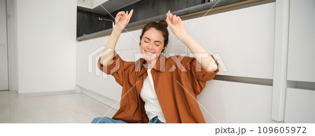 Dancing girl sits on kitchen floor, moving to the rhythm, enjoying sound quality in wireless headphones, listening to music in earphones Dancing girl sits on kitchen floor, moving to the rhythm, enjoying sound quality in wireless headphones, listening to music in earphones 109605972