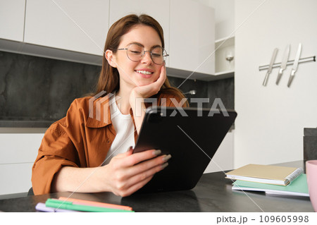 E-learning and remote workplace concept. Young woman in glasses, student studying at home in kitchen, reading on digital tablet, looking at her project, connects to online meeting E-learning and remote workplace concept. Young woman in glasses, student studying at home in kitchen, reading on digital tablet, looking at her project, connects to online meeting 109605998
