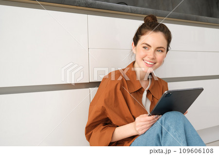 Portrait of young beautiful woman sitting on kitchen floor with digital tablet, browsing news feed, social media app on gadget, smiling and looking happy Portrait of young beautiful woman sitting on kitchen floor with digital tablet, browsing news feed, social media app on gadget, smiling and looking happy 109606108