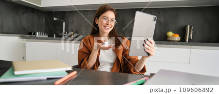 Portrait of young woman working from home, sitting in kitchen and talking with team, has online meeting, using digital tablet to join webinar, having conversation 109606892