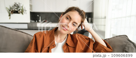 Close up portrait of young woman resting at home, girl sits on sofa, smiles at camera, enjoys her weekend indoors, watches television Close up portrait of young woman resting at home, girl sits on sofa, smiles at camera, enjoys her weekend indoors, watches television 109606899