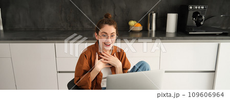 Portrait of beautiful woman in glasses, sits at home in kitchen, looks at computer screen with amazed, surprised face, reacts to big great news seeing it on laptop 109606964