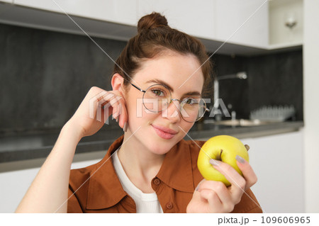 Beautiful young woman in glasses, sitting on chair in the kitchen, eating green apple and smiling 109606965