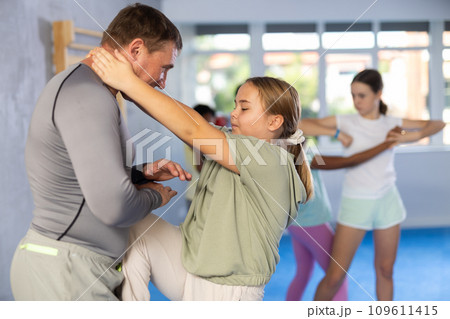 Tween girl performing knee strike in midsection to instructor in self-defense sparring 109611415