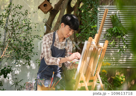 Young happy Asian man artist in an apron is coloring on a canvas on an easel in his home garden. 109613718
