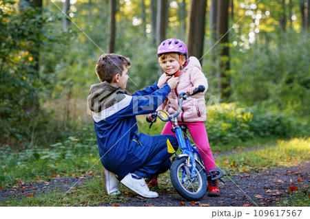 School kid boy, brother put on little preschool sister girl bike helmet on head. Brother teaching happy child cycling and having fun with learning bike. Active siblings family outdoors. Kids activity 109617567