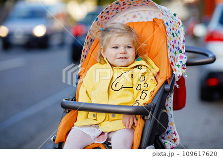 Portrait of little cute toddler girl sitting in stroller or pram and going for a walk. Happy cute baby child having fun outdoors. Healthy daughter. Street traffic in the city. 109617620