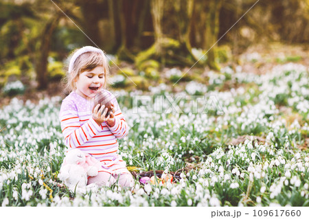 Little girl with Easter bunny ears making egg hunt in spring forest on sunny day, outdoors. Cute happy child with lots of snowdrop flowers, huge chocolate egg and colored eggs. 109617660
