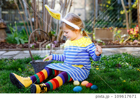 Beautiful funny little toddler girl with bunny ears having fun with traditional Easter eggs hunt on sunny day, outdoors. Happy child celebrating holiday. Baby find and collect colored eggs in garden Beautiful funny little toddler girl with bunny ears having fun with traditional Easter eggs hunt on sunny day, outdoors. Happy child celebrating holiday. Baby find and collect colored eggs in garden 109617671