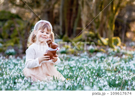Happy little girl with Easter bunny ears eating chocolate figure in spring forest on sunny day, outdoors. Cute child with lots of snowdrop flowers. Springtime, christian holiday concept. 109617674