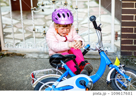Cute little girl sitting on the ground after falling off her bike. Upset crying preschool child with safe helmet getting hurt while riding a bicycle. Active family leisure with kids. Cute little girl sitting on the ground after falling off her bike. Upset crying preschool child with safe helmet getting hurt while riding a bicycle. Active family leisure with kids. 109617681