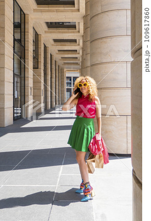Content fashion female with bag walking on pavement near stone columns in sunlight and talk cell phone.  109617860