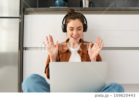 Portrait of happy young woman, freelancer working from home, remote worker sits on floor with laptop and headphones, looks surprised and excited at screen Portrait of happy young woman, freelancer working from home, remote worker sits on floor with laptop and headphones, looks surprised and excited at screen 109620443