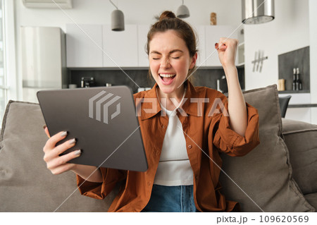 Portrait of excited young woman with digital tablet, sitting on couch, laughing and smiling, winning on video game, spending time at home 109620569