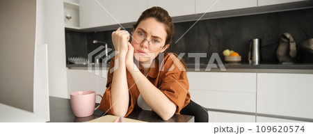 Portrait of sad and tired young woman in glasses, sitting in kitchen at home, looking upset and exhausted, working from home Portrait of sad and tired young woman in glasses, sitting in kitchen at home, looking upset and exhausted, working from home 109620574