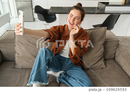 Portrait of beautiful happy girl takes selfies on sofa. Woman posing for photos on her couch, taking pictures for social media, extends hand with smartphone Portrait of beautiful happy girl takes selfies on sofa. Woman posing for photos on her couch, taking pictures for social media, extends hand with smartphone 109620593
