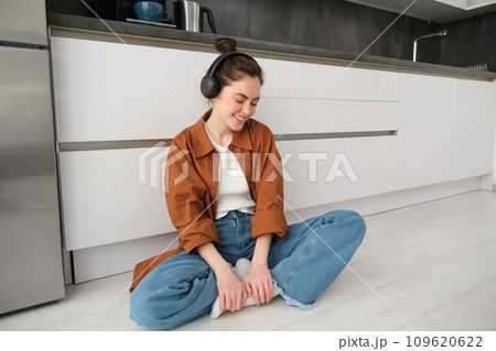 Cute modern girl on kitchen floor at home, listening music in wireless headphones, having fun 109620622