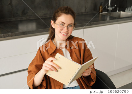 Portrait of young self-employed freelance tutor, sitting in kitchen at home, holding notebook with study notes, revising for university Portrait of young self-employed freelance tutor, sitting in kitchen at home, holding notebook with study notes, revising for university 109620799