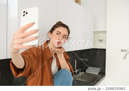 Beautiful young woman, social media influencer takes pictures, records video with smartphone camera, posing on kitchen counter at home, lifestyle blog and people concept Beautiful young woman, social media influencer takes pictures, records video with smartphone camera, posing on kitchen counter at home, lifestyle blog and people concept 109620962