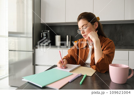 Portrait of young woman student, girl studying at home, working remote in kitchen, taking notes, sitting indoors 109621004