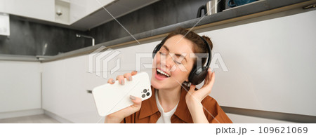 Portrait of happy, beautiful girl sings songs in headphones, listens to music, using smartphone as mic, playing karaoke app Portrait of happy, beautiful girl sings songs in headphones, listens to music, using smartphone as mic, playing karaoke app 109621069
