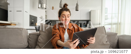 Portrait of anxious, stressed young woman looking scared at digital tablet, staring at screen, sitting on couch with gadget in living room Portrait of anxious, stressed young woman looking scared at digital tablet, staring at screen, sitting on couch with gadget in living room 109621077