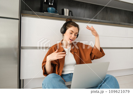 Portrait of woman listens to music in headphones, while working on laptop, sits at home on kitchen floor, making dance movements 109621079