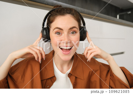 Excited woman likes sound quality, points at her wireless headphones and smiles, sits in kitchen on floor and looks happy Excited woman likes sound quality, points at her wireless headphones and smiles, sits in kitchen on floor and looks happy 109621085