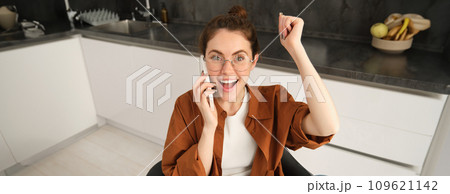 Portrait of woman receiving great news over the phone. Girl talking on mobile telephone and celebrating, laughing and making fist pump, dancing on her chair in kitchen Portrait of woman receiving great news over the phone. Girl talking on mobile telephone and celebrating, laughing and making fist pump, dancing on her chair in kitchen 109621142