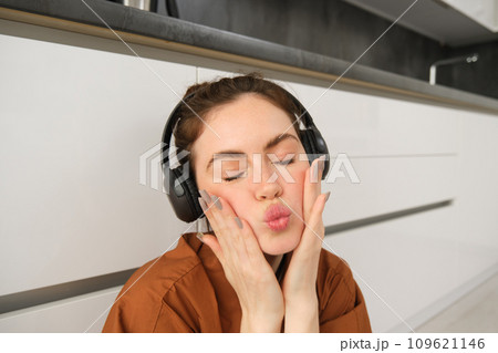 Beautiful young woman in wireless headphones, makes silly and cute faces, listens to music at home 109621146