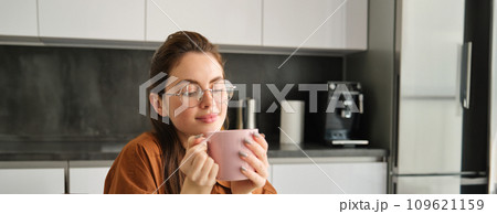 Portrait of beautiful young woman, smells aroma of her coffee in mug, drinks cup, sits in kitchen Portrait of beautiful young woman, smells aroma of her coffee in mug, drinks cup, sits in kitchen 109621159