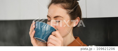Portrait of beautiful young woman, enjoying delicious aroma of freshly made coffee, holding mug, standing near machine in the kitchen, drinking from mug Portrait of beautiful young woman, enjoying delicious aroma of freshly made coffee, holding mug, standing near machine in the kitchen, drinking from mug 109621206