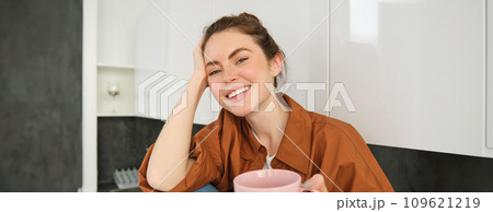 Beautiful woman offers you her cup of coffee, smiling and looking happy, sitting in kitchen, enjoying her morning drink Beautiful woman offers you her cup of coffee, smiling and looking happy, sitting in kitchen, enjoying her morning drink 109621219