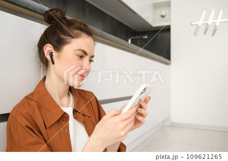 Close up portrait of young modern woman sits on kitchen floor, looking at her smartphone, watching video or tv series on streaming service app, listening music in wireless headphones Close up portrait of young modern woman sits on kitchen floor, looking at her smartphone, watching video or tv series on streaming service app, listening music in wireless headphones 109621265