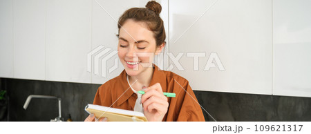Portrait of smiling young woman with notebook, writing, making notes in planner, sitting at home in the kitchen 109621317