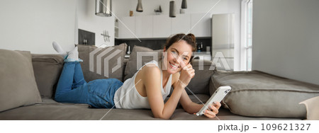 Portrait of cute, happy young woman lying on couch, holding mobile phone, using smartphone while relaxing at home in living room, concept of lifestyle and people leisure Portrait of cute, happy young woman lying on couch, holding mobile phone, using smartphone while relaxing at home in living room, concept of lifestyle and people leisure 109621327