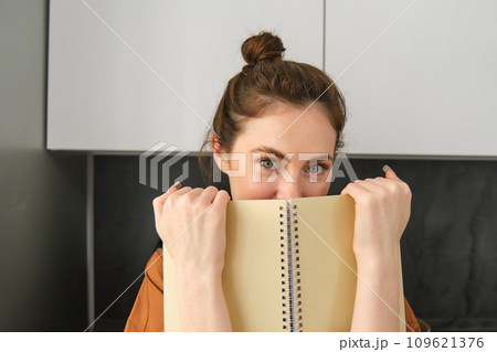 Lifestyle and people concept. Young brunette woman, holding notebook near face, hiding smile behind notepad and looking coquettish at camera Lifestyle and people concept. Young brunette woman, holding notebook near face, hiding smile behind notepad and looking coquettish at camera 109621376
