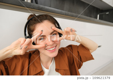 Close up of carefree young modern woman, relaxes at home, listens music in wireless headphones, shows v-sign and smiles at camera 109621379