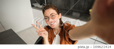 Selfie of cute young woman in glasses, posing at home in kitchen, taking pictures on her mobile phone, smiling and showing peace sign 109621414