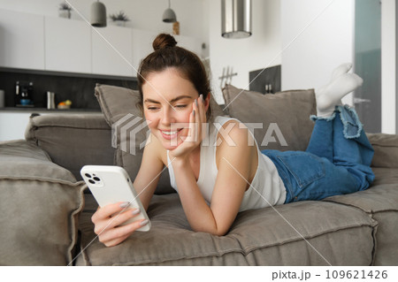 Portrait of smiling, happy young woman resting at home, resting on couch with smartphone, using mobile phone application 109621426