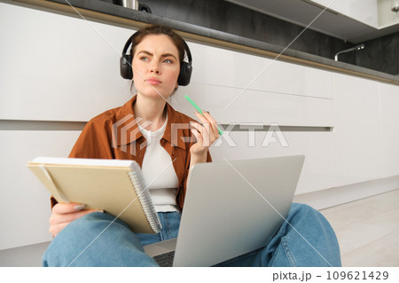 Portrait of young woman, modern student sits at home on floor and doing homework on laptop, listens music in headphones while making assignment or project for college 109621429