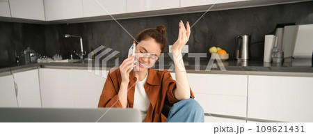 Excited young woman talking on mobile phone in front of laptop, sitting in kitchen with happy face expression, having a conversation 109621431