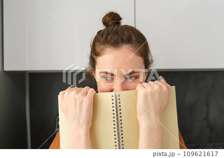 Cute woman hides face behind notebook, looking aside, smiling with eyes Cute woman hides face behind notebook, looking aside, smiling with eyes 109621617