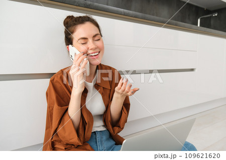 Carefree girl laughs, talks on mobile phone, calling friend and having funny conversation, sitting casually on kitchen floor 109621620