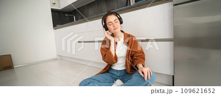 Technology concept. Young smiling brunette woman in wireless headphones, listens to music, sits on kitchen floor Technology concept. Young smiling brunette woman in wireless headphones, listens to music, sits on kitchen floor 109621652