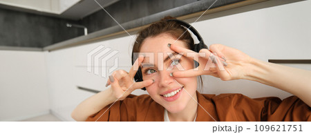 Close up portrait of woman in headphones, shows peace, v-signs near eyes, smiles and looks happy, sits on floor at home 109621751