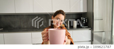 Beautiful young woman in glasses, giving you cup of tea, passing mug with aromatic delicious coffee, sitting in kitchen at home 109621787