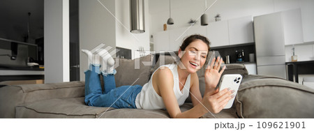 Happy young woman video chatting with friends, using smartphone app, waving hand at smartphone screen and saying hello Happy young woman video chatting with friends, using smartphone app, waving hand at smartphone screen and saying hello 109621901