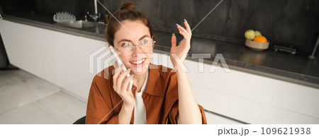 Excited young woman talking on mobile phone in front of laptop, sitting in kitchen with happy face expression, having a conversation 109621938